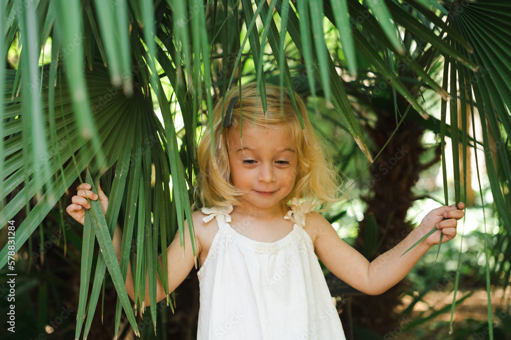 Cute caucasian pretty blonde two year old girl, toddler, kid hiding among big green exotic leaves in botanic garden. Natural beauty,perfect skin, health,cosmetics concept. Cute caucasian pretty blonde two year old girl, toddler, kid hiding among big green exotic leaves in botanic garden. Natural beauty,perfect skin, health,cosmetics concept.