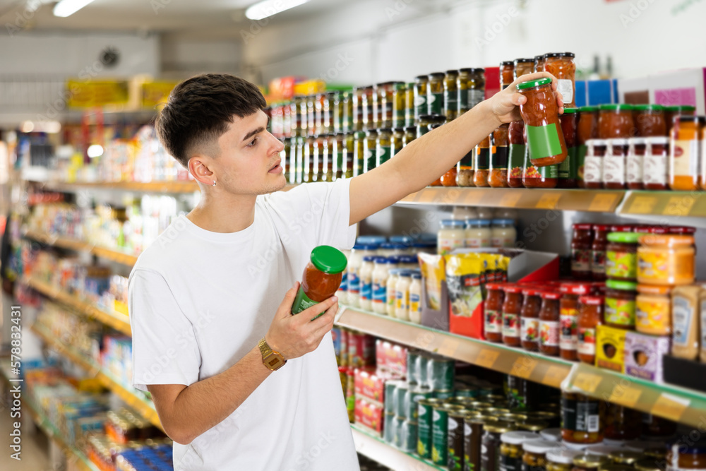 Focused thoughtful young guy choosing food in supermarket, holding glass jars with canned goods and reading labels