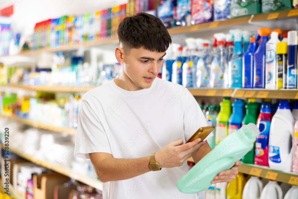 Focused young guy scanning barcode on plastic bottle of household detergent with smartphone while shopping in supermarket, paying for item using mobile app