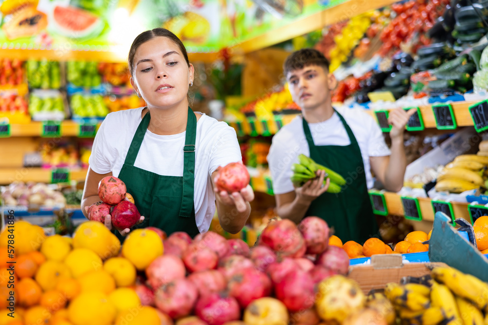 Confident young saleswoman working in a vegetable store puts pomegranates on the counter for sale