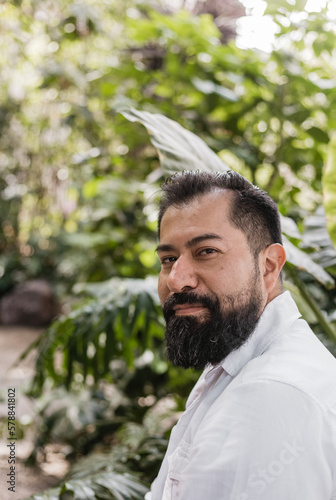Retrato de hombre mexicano en edad adulta , con barba, plantas en su entorno, camisa blanca y pantalón beige