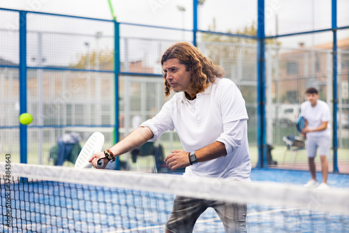 Portrait of middle-aged Latin male in sportswear enjoying popular sport padel game on tennis court outside