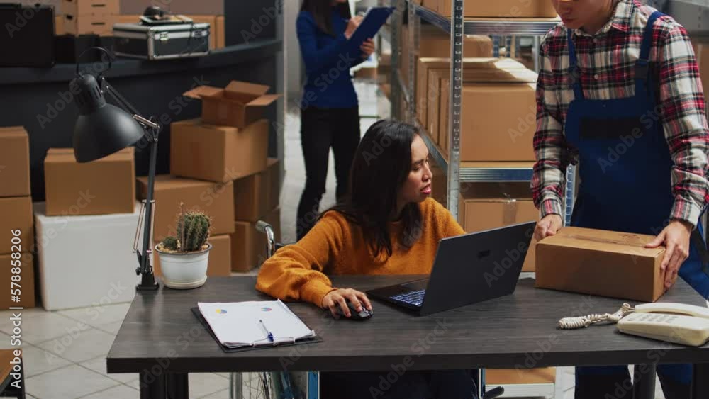 Young employee with impairment working in warehouse, doing desk work for stock logistics and inventory on laptop. Woman wheelchair user doing financial planning in disability friendly space.