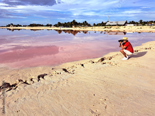Latin adult photographer man with shorts  red shirt and hat takes pictures on the sand next to the pink colored lagoon with a high concentration of salt  Las Coloradas in Yucatan Mexico