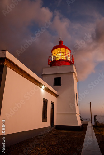 The classic Illuminated lighthouse with a lamp on during the sunset, dramatic clouds in the sky.
