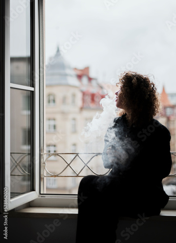 Young beautiful woman with curly hair smoking cigarettes near the open window © Alona