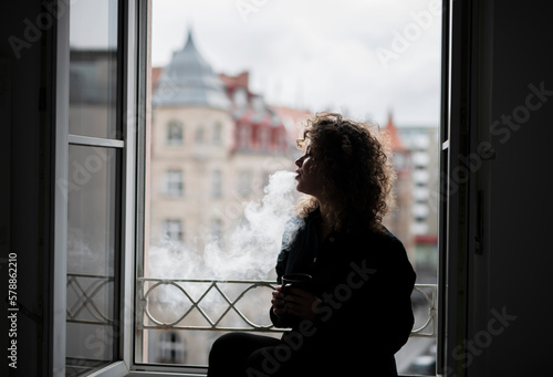 Young beautiful woman with curly hair smoking cigarettes near the open window © Alona