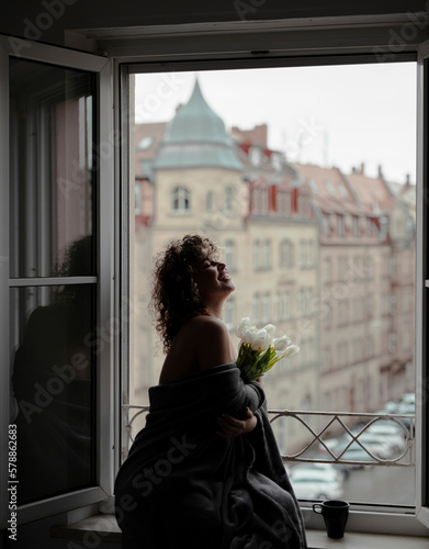Young beautiful woman with curly hair standing near the open window, with bouquet of tulips in the morning © Alona