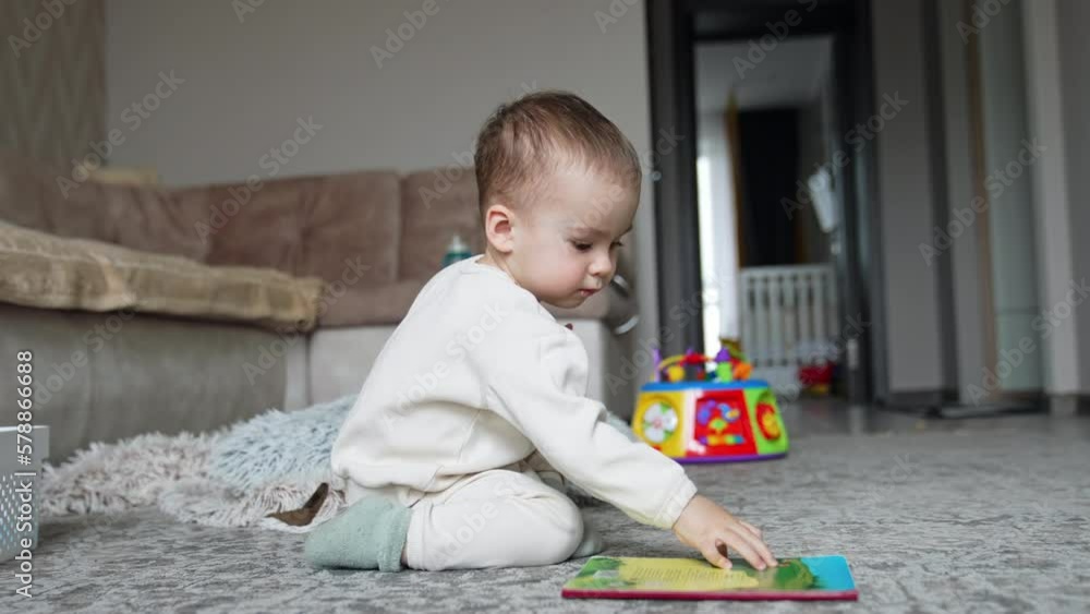 Lovely toddler in white clothes sitting on the floor in the room. Baby boy looking at book and eating a cookie. Lovely toddler in white clothes sitting on the floor in the room. Baby boy looking at book and eating a cookie.