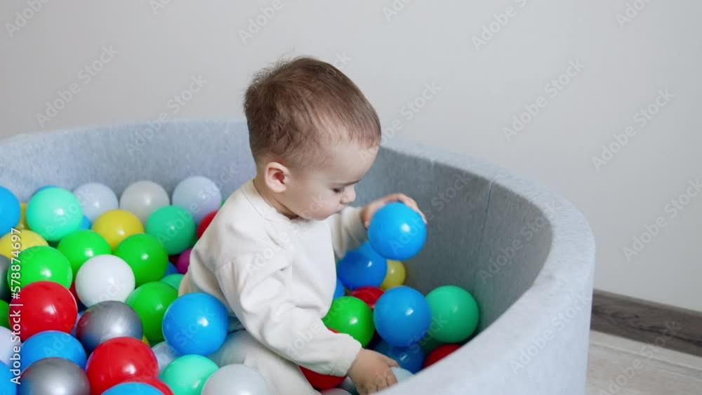 Cute Caucasian baby playing in the dry basin. Kid in white sport suit throws ball out of basin and lies back on the toys. Cute Caucasian baby playing in the dry basin. Kid in white sport suit throws ball out of basin and lies back on the toys.