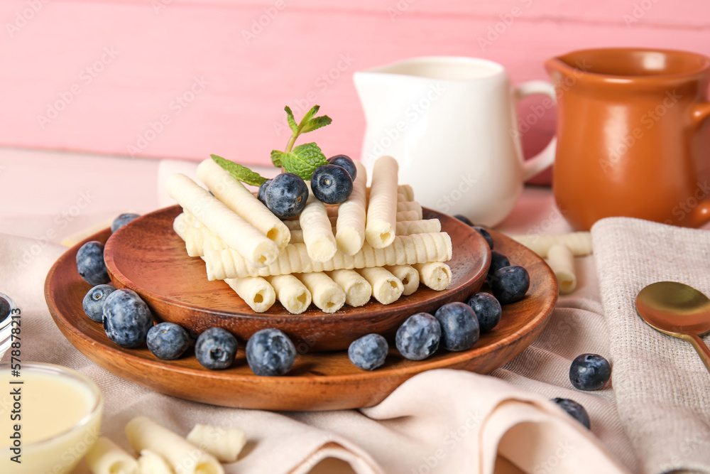 Plates with delicious wafer rolls and blueberries on white wooden table against pink background Plates with delicious wafer rolls and blueberries on white wooden table against pink background
