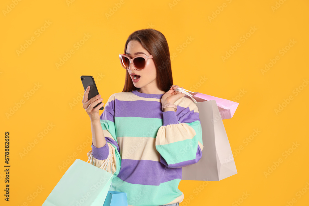 Surprised young woman with shopping bags looking at smartphone on yellow background. Big sale Surprised young woman with shopping bags looking at smartphone on yellow background. Big sale