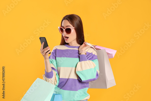 Surprised young woman with shopping bags looking at smartphone on yellow background. Big sale