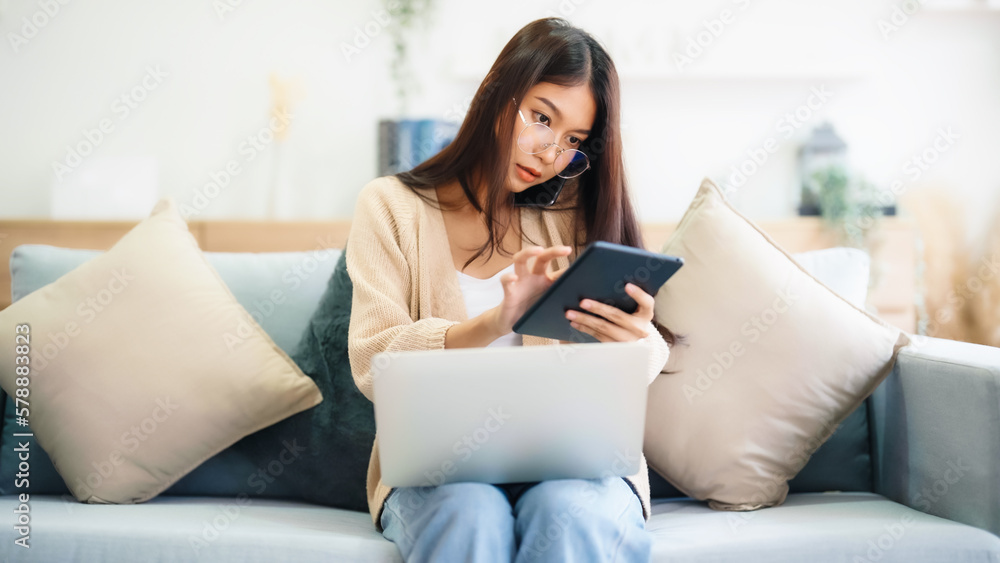 Young asian woman using computer laptop and mobile smartphone while seated on couch at home Young asian woman using computer laptop and mobile smartphone while seated on couch at home