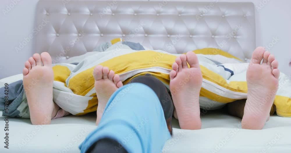 Dachshund dog jumps out from under blanket between bare feet of young couple. Man and woman