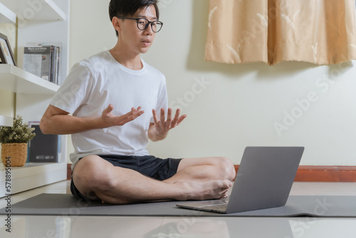 Asian healthy trainer man using laptop for teaching online healthcare yoga and meditation course at home. Man teaching breathing for meditating and relief stress. Online healthcare at home concept.