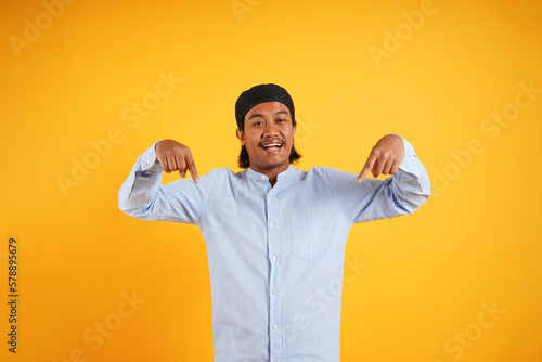 Photo of excited asian young moslem man with two fingers pointing down, isolated on yellow background photo