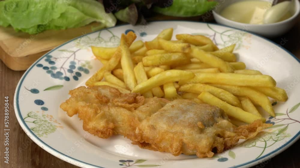 Using knife spreading homemade sauce on crispy delicious battered fish with pile of french fried in white ceramic dish on wooden table. Using knife spreading homemade sauce on crispy delicious battered fish with pile of french fried in white ceramic dish on wooden table.