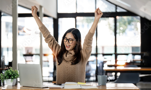 Elegant businesswoman sitting in office with digital tablet. Excited asian businesswoman raising hands to congratulate while working on laptop in office.