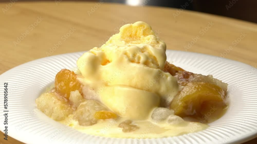 Using metal spoon scooping bite of delicious homemade peach cobbler topping with vanilla ice cream in white ceramic plate on wooden table. Using metal spoon scooping bite of delicious homemade peach cobbler topping with vanilla ice cream in white ceramic plate on wooden table.