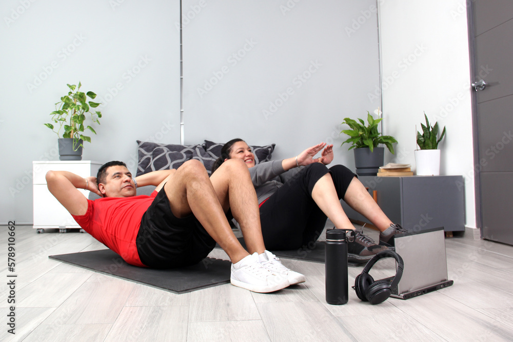 Obraz premium Latino adult couple of man and woman exercise at home taking online class they see the laptop in their living room together they exercise to avoid diabetes and hypertension