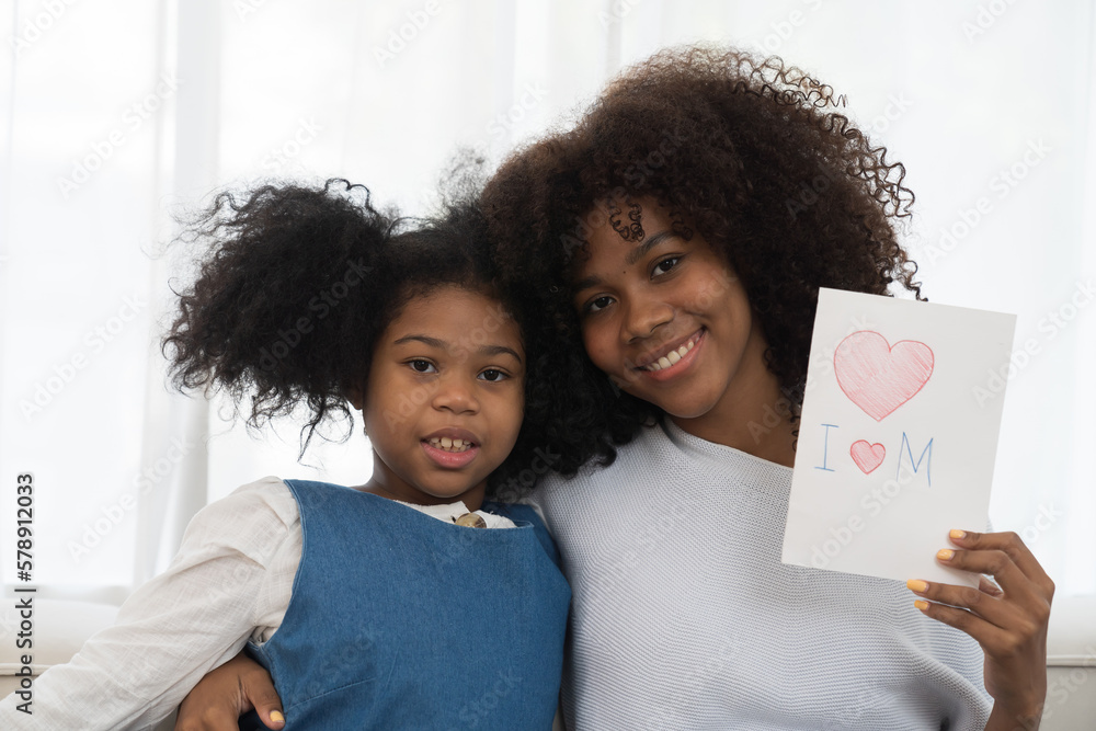 Happy family embracing together on sofa and holding mother day paper card at home. Smiling African American young mother spending time with her child daughter at home Happy family embracing together on sofa and holding mother day paper card at home. Smiling African American young mother spending time with her child daughter at home