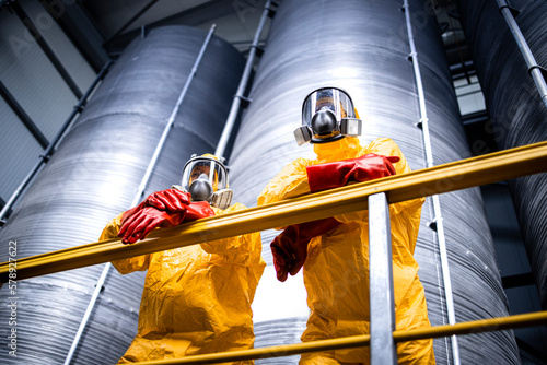 Industrial factory interior with workers in protection suit, gas mask, gloves standing by large storage tanks or reservoirs with chemicals.