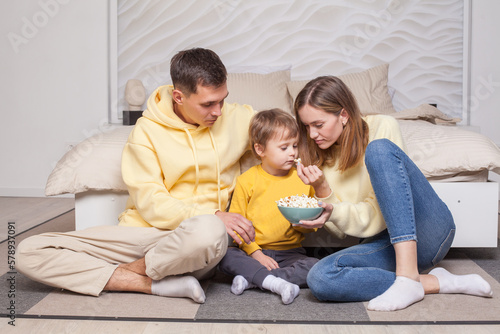 Friendly family, lovely parents in yellow clothes with child son sitting by the bed and holding bowl with popcorn