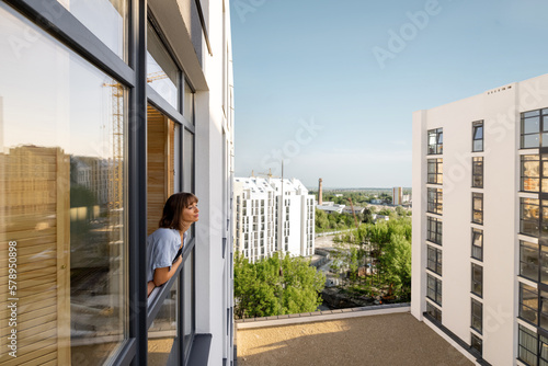 Woman looks out the window of apartment building in the city, urban top view from the outside, sunny weather. Concept of living in apartment building, quarantine and loneliness