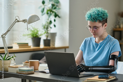 Girl with prosthetic arm working on laptop at her workplace in the room, she having remote work photo