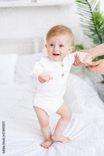 mom's hands are holding a baby 6 months old, a blond boy with blue eyes on a white bed in a light bedroom in a white cotton bodysuit, the concept of children's goods