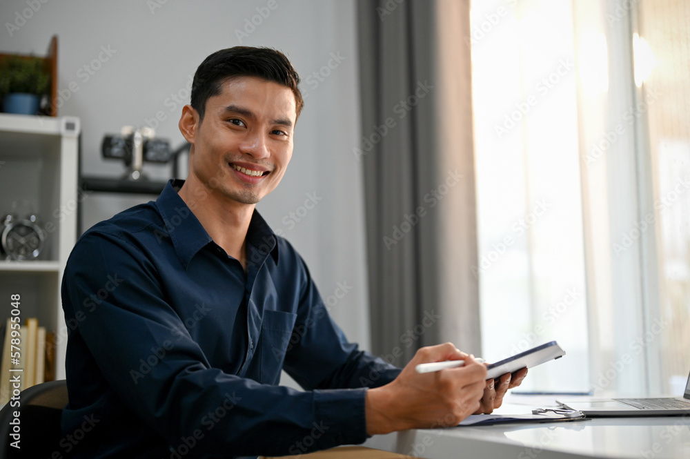 Successful Asian businessman holding a tablet, sitting at his desk, smiling and looking at the camera. Successful Asian businessman holding a tablet, sitting at his desk, smiling and looking at the camera.