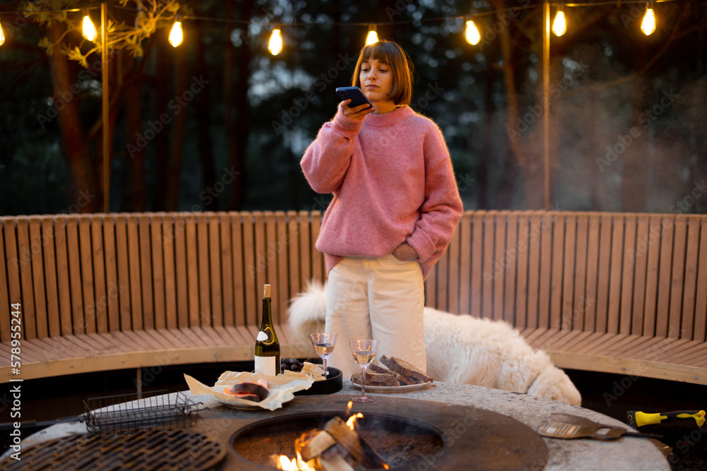 Fototapeta premium Young woman spends leisure time with her dog, taking photo on phone at beautiful lounge area with bonfire and round bench in pine forest at dusk