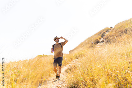 Caucasian man backpacker hiking on mountain hill in sunny day. Handsome guy enjoy adventure outdoor lifestyle walking on mountain dirt road on summer vacation. Ecotourism and solo travel concept.