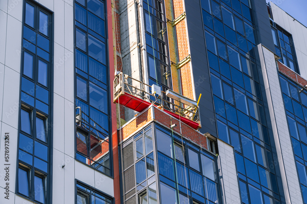 Construction lift for finishing works at height. A construction worker works in a cradle of a lifting mechanism at a height. High-rise works during the construction of a residential building. Construction lift for finishing works at height. A construction worker works in a cradle of a lifting mechanism at a height. High-rise works during the construction of a residential building.