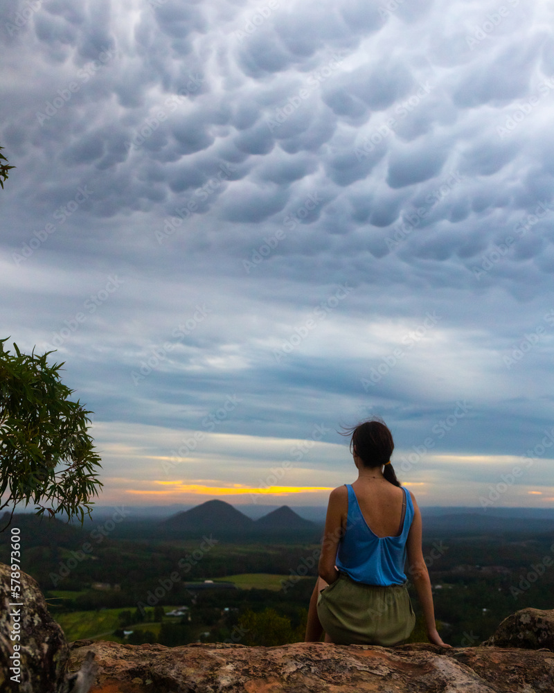 A girl sits on top of Mount Ngungun and admires a rare phenomenon, the spectacular Mammatus clouds formed during sunset in Glass House Mountains National Park, Queensland, Australia