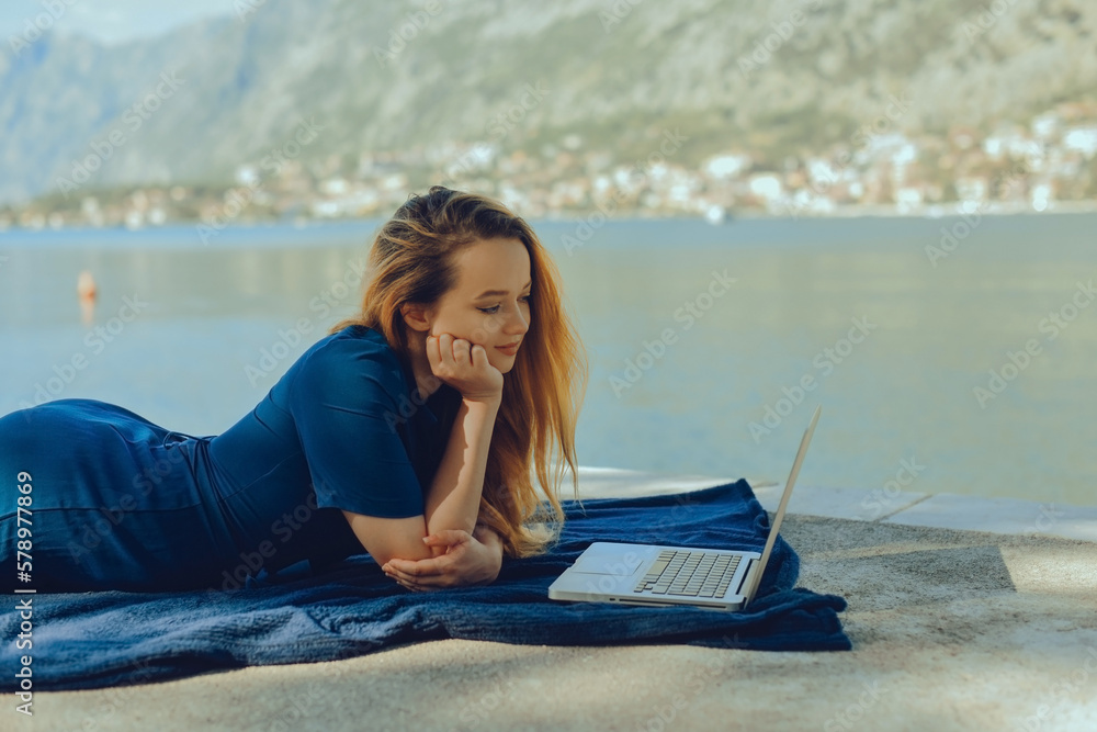 Girl freelancer lies on the background of the mountains with a laptop, remote work. A woman looks at the screen and writes something on the beach in the summer.