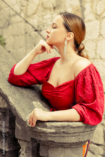 A beautiful girl in red is standing on the old stairs to the castle. Woman in accessories and professional make-up. Fashion and style © MoreThanProd