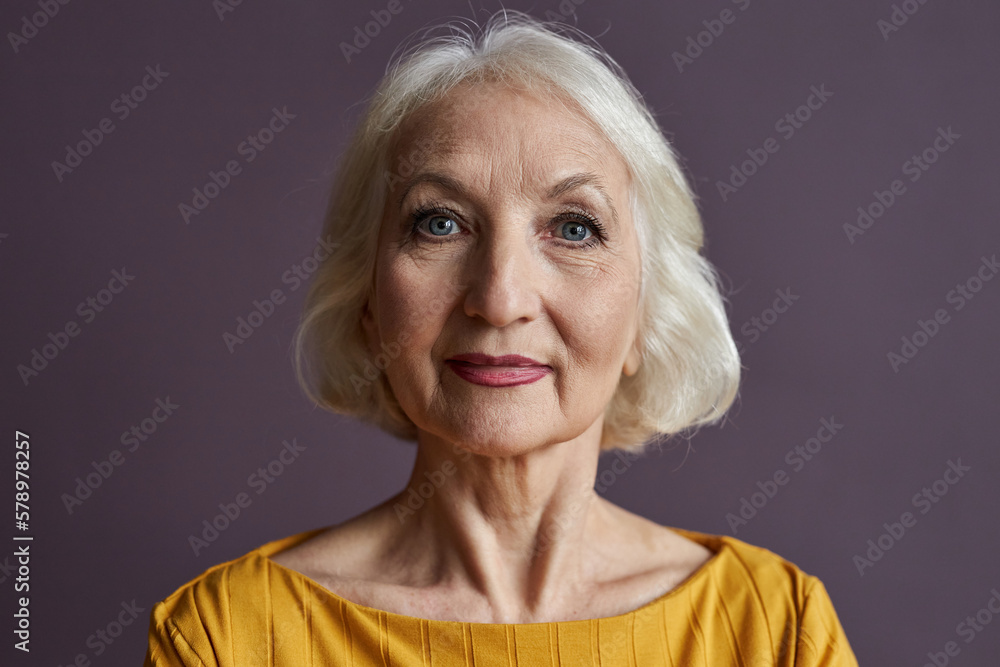 Minimal close up portrait of of elegant senior woman wearing makeup and looking at camera against dusty purple background Minimal close up portrait of of elegant senior woman wearing makeup and looking at camera against dusty purple background