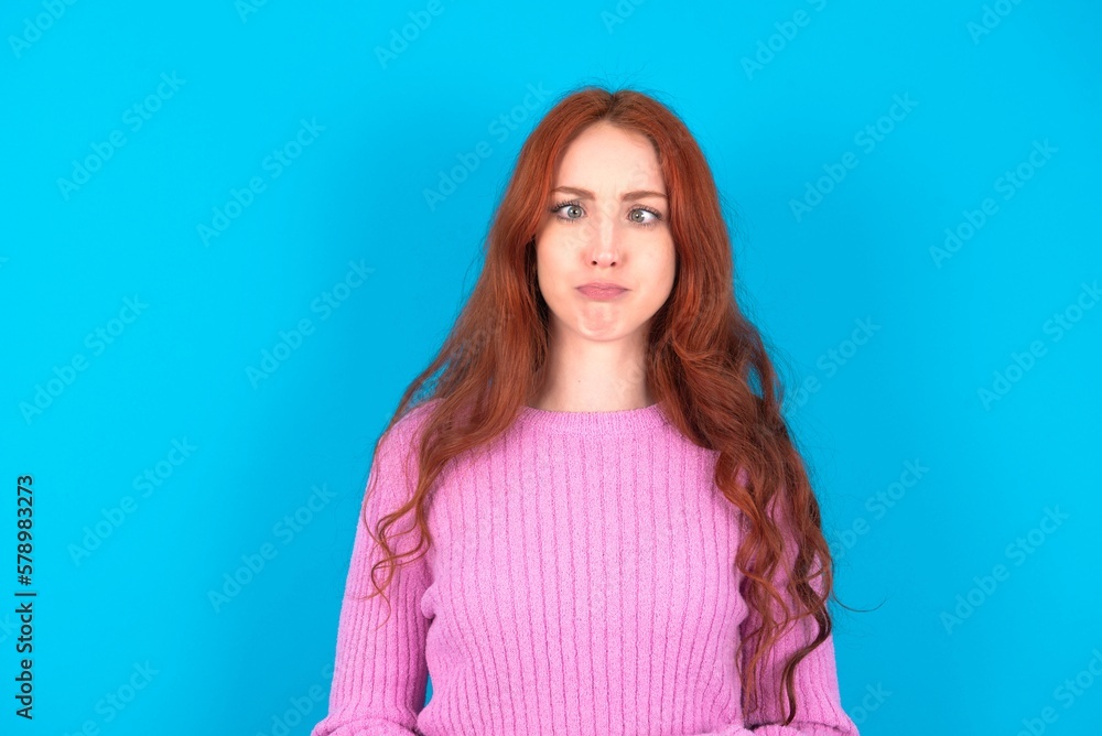 Fototapeta premium young woman wearing pink sweater over blue background crosses eyes, puts lips, makes grimace with awkward expression has fun alone, plays fool.
