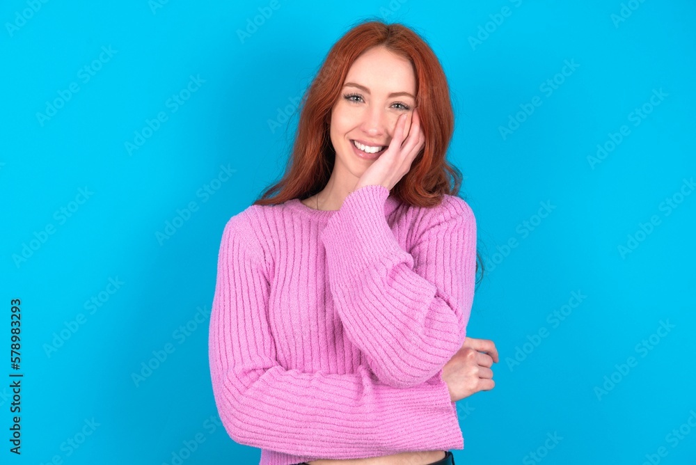 Shocked, astonished young woman wearing pink sweater over blue background looking surprised in full disbelief wide open mouth with hand near face. Positive emotion facial expression body language. Shocked, astonished young woman wearing pink sweater over blue background looking surprised in full disbelief wide open mouth with hand near face. Positive emotion facial expression body language.