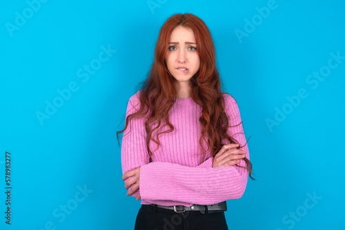 young woman wearing pink sweater over blue background bitting his mouth and looking worried and scared crossing arms, worry and doubt.