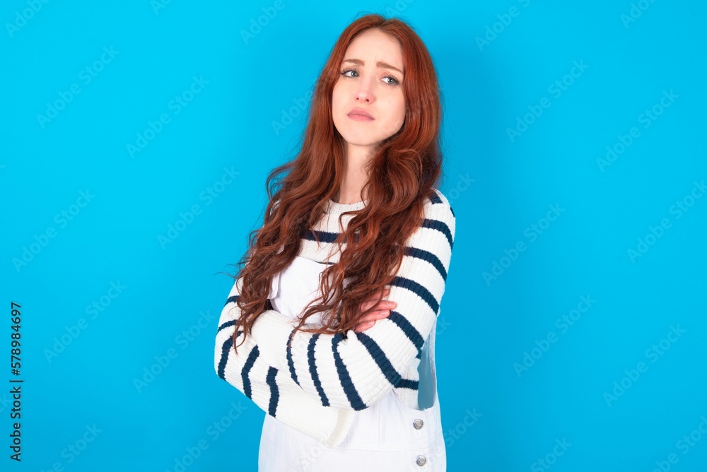Image of upset young caucasian woman wearing overalls over blue background with arms crossed. Looking with disappointed expression aside after listening to bad news. Image of upset young caucasian woman wearing overalls over blue background with arms crossed. Looking with disappointed expression aside after listening to bad news.
