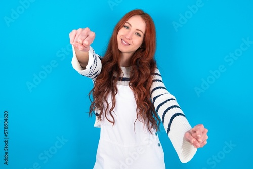 Portrait of charming young caucasian woman wearing overalls over blue background , smiling broadly while holding hands over her head. Confidence and relax concept.