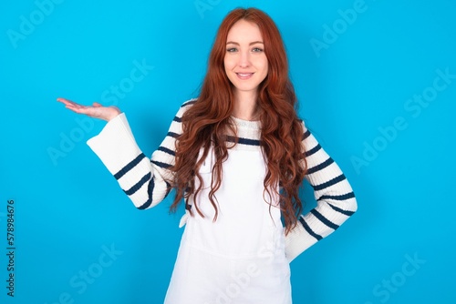 Positive glad young caucasian woman wearing overalls over blue background says: wow how exciting it is, has amazed expression, shows something on blank space with open hand. Advertisement concept.