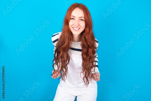 young caucasian woman wearing overalls over blue background raising fists up screaming with joy being happy to achieve goals.