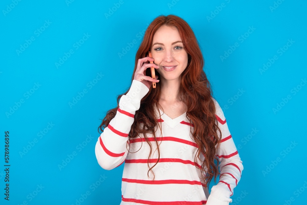 Portrait of a smiling young woman wearing striped sweater over blue background talking on mobile phone. Business, confidence and communication concept. Portrait of a smiling young woman wearing striped sweater over blue background talking on mobile phone. Business, confidence and communication concept.
