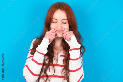 Pleased young woman wearing striped sweater over blue background with closed eyes keeps hands near cheeks and smiles tenderly imagines something very pleasant