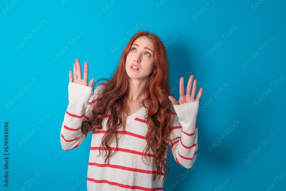 young woman wearing striped sweater over blue background keeps palms forward and looks with fright above on ceiling tries to defense herself from invisible danger opens mouth. young woman wearing striped sweater over blue background keeps palms forward and looks with fright above on ceiling tries to defense herself from invisible danger opens mouth.