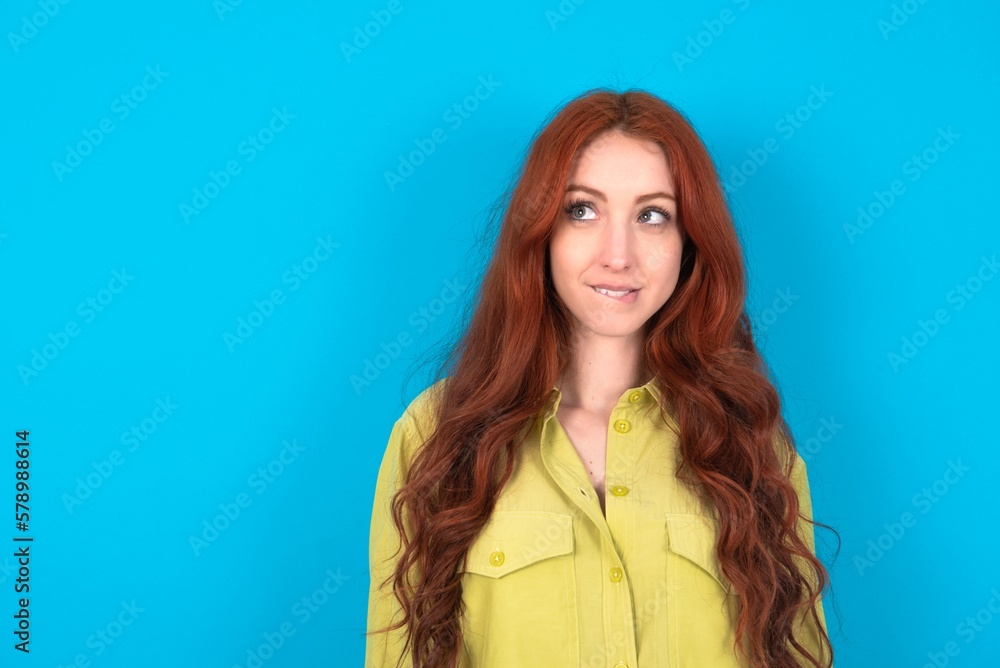 Amazed young woman wearing green sweater over blue background bitting lip and looking tricky to empty space. Amazed young woman wearing green sweater over blue background bitting lip and looking tricky to empty space.
